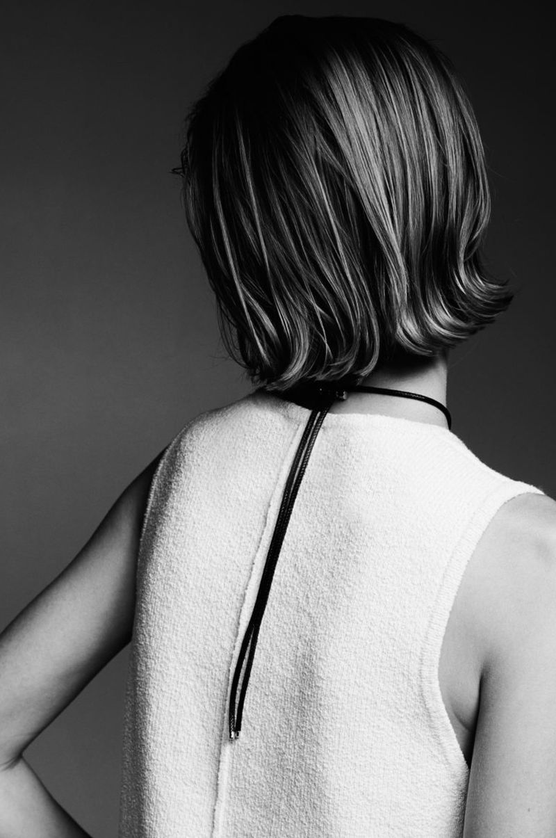 Back view of a woman with blonde bob hairstyle, wearing a white sleeveless top and a black choker necklace, against a grey backdrop.