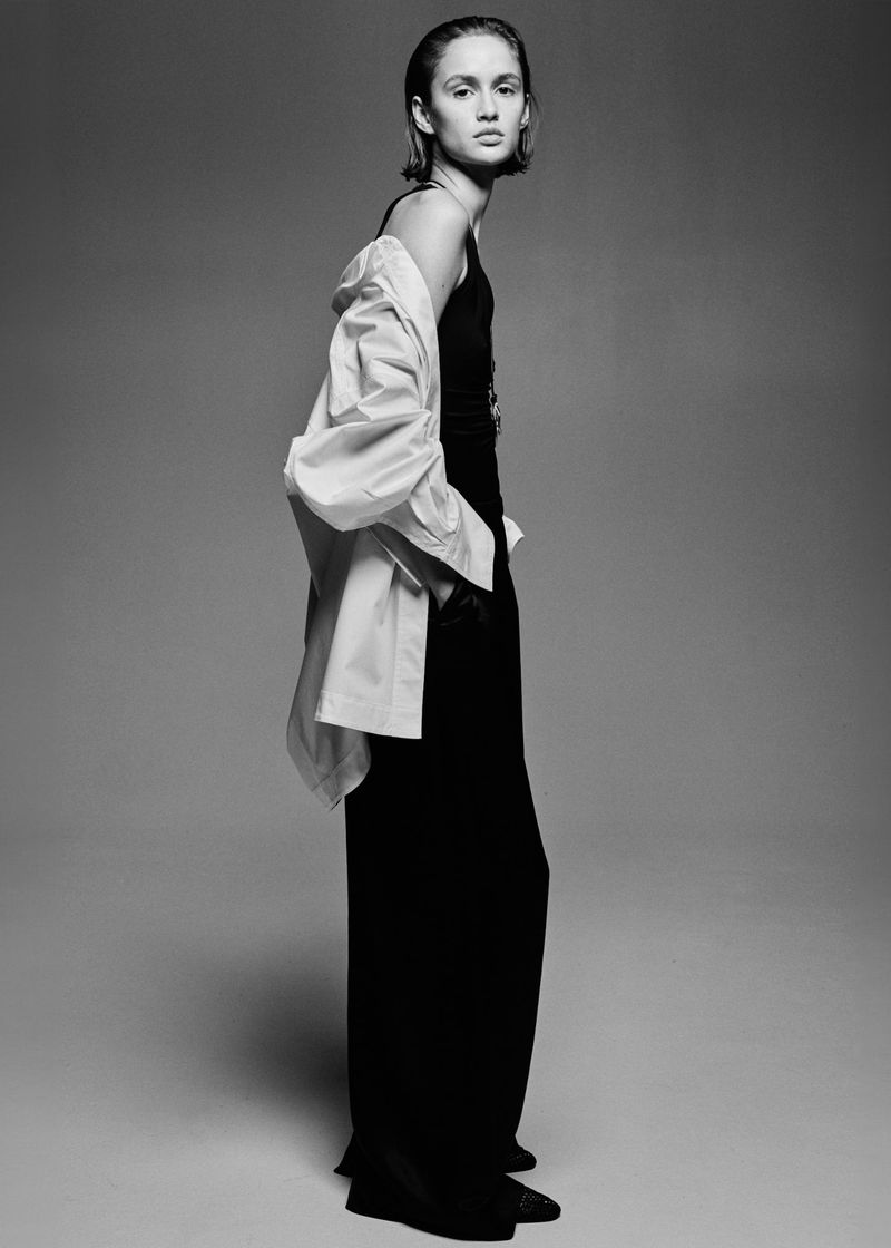 Young woman in black tank top and wide-legged pants poses in photo studio wearing white button-down shirt, standing tall looking forward.