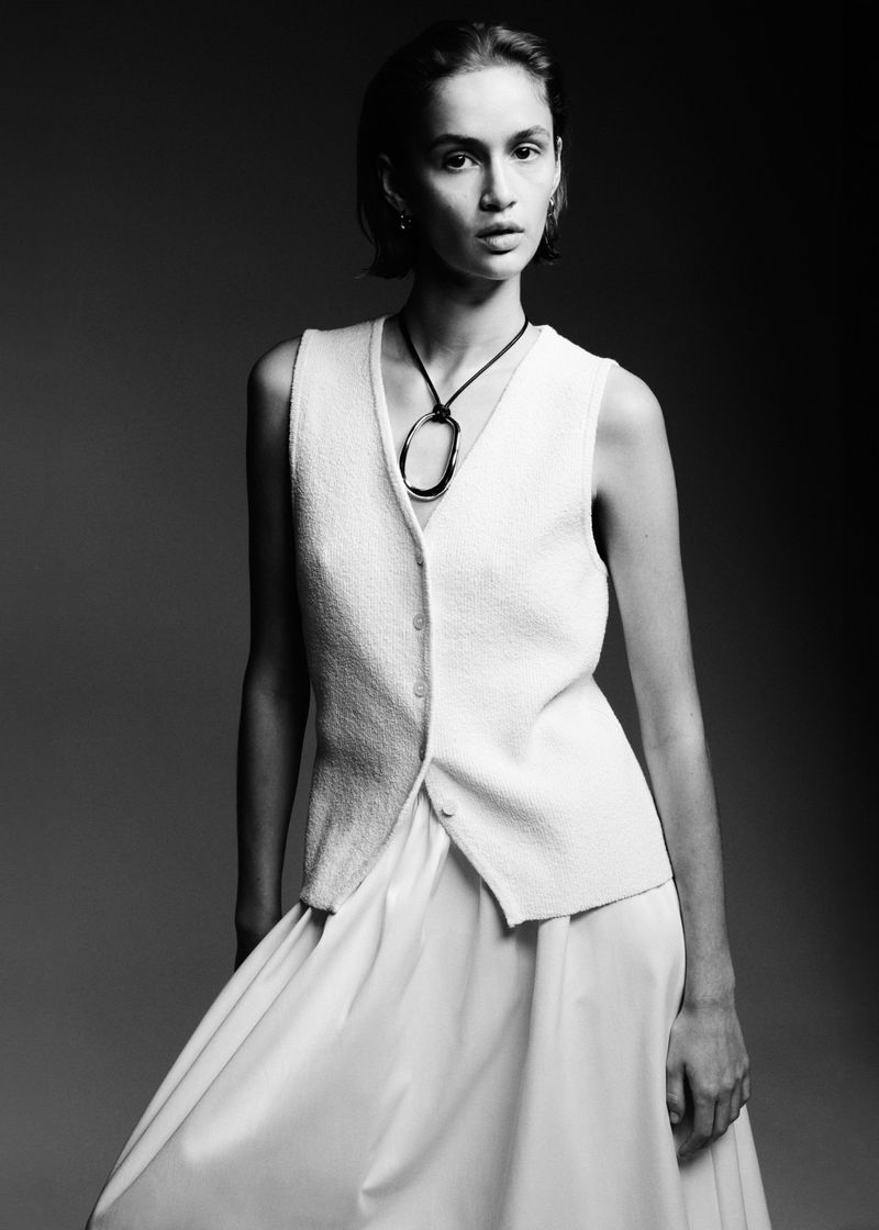 Elegant portrait of a woman in a white vest and skirt, accessorized with a striking black pendant necklace, in a studio setting.