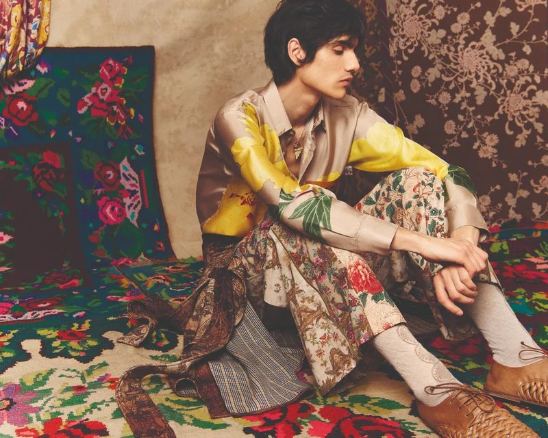 Male fashion model sits cross-legged on a floral rug. He wears a yellow and tan floral shirt and pants, with a matching backdrop.