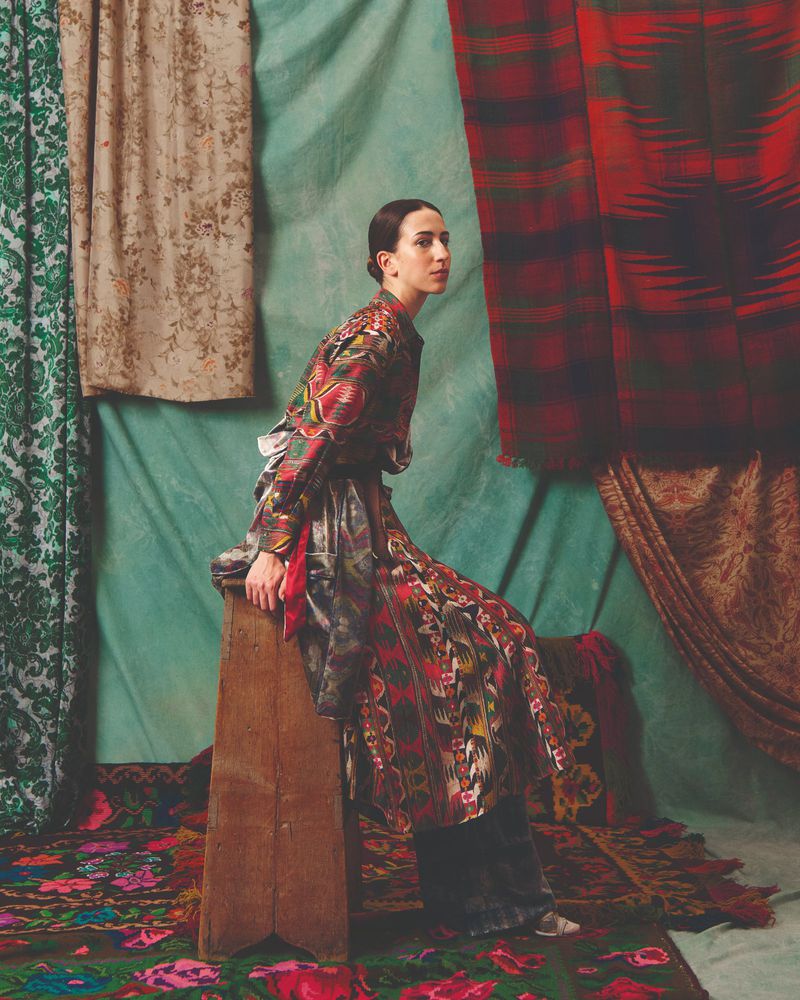 An adult female model sits on a wooden stool in front of a backdrop with layered textile patterns, wearing a patterned, long-sleeve dress.
