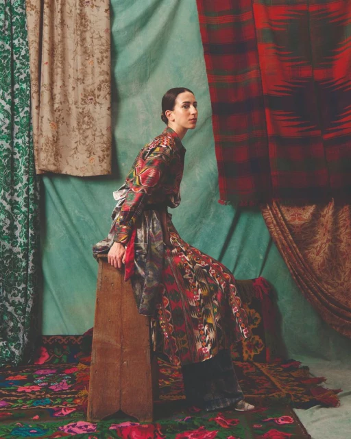 An adult female model sits on a wooden stool in front of a backdrop with layered textile patterns, wearing a patterned, long-sleeve dress.