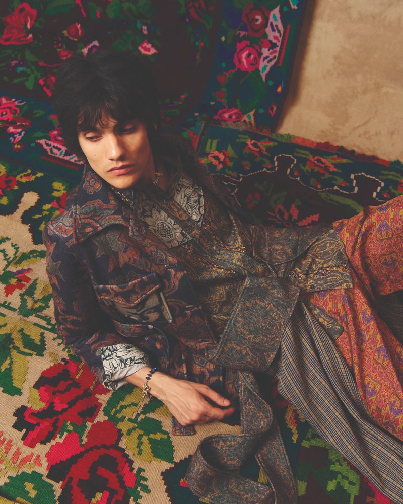 A stylish adult male with dark hair lies on a floral patterned rug wearing a patterned jacket and pants in a close-up studio shot.