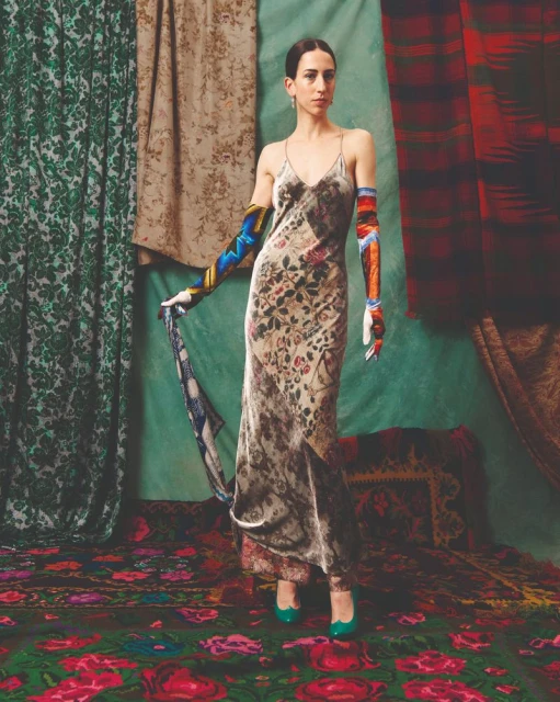 A woman poses in a patterned dress with long, colorful gloves against a backdrop of draped patterned fabrics, with matching patterned carpet.