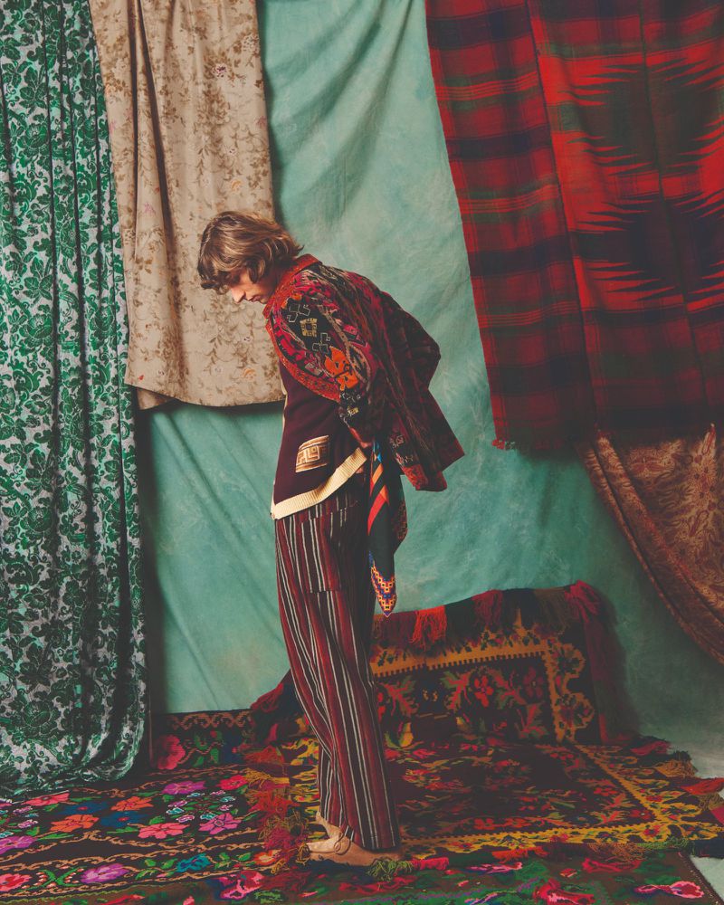 A person in patterned clothing stands amid patterned textiles in a studio with green and red hues, embodying vintage and fashion elements.