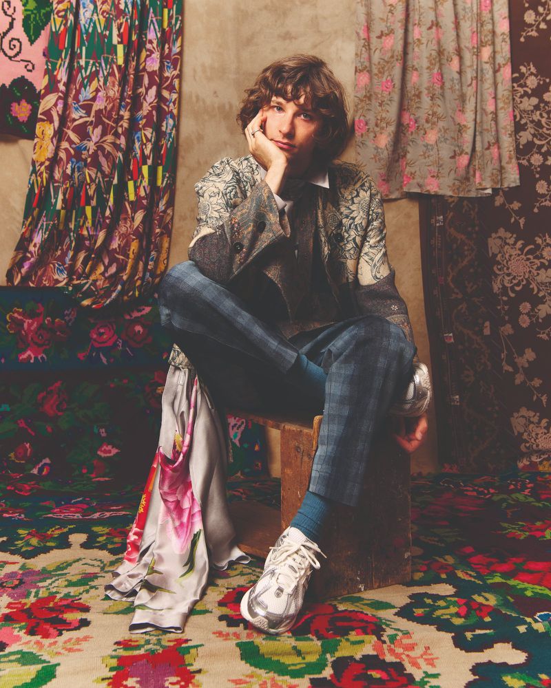 Adult male seated on a wooden stool, wearing a patterned blazer with blue plaid pants, set against fabric backdrops.