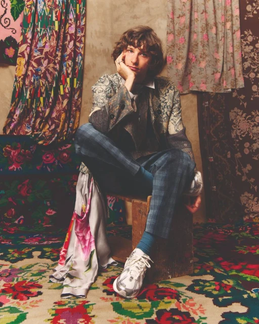 Adult male seated on a wooden stool, wearing a patterned blazer with blue plaid pants, set against fabric backdrops.