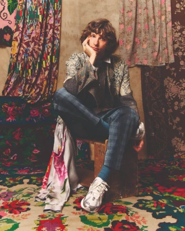 Adult male seated on a wooden stool, wearing a patterned blazer with blue plaid pants, set against fabric backdrops.