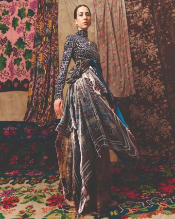 An adult female is dressed in patterned garments, posing in a photo studio with a patterned carpet and draped fabric backdrop.