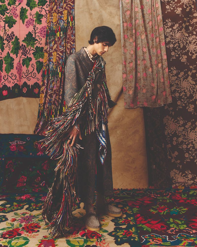 A young adult poses in a fashion editorial wearing a grey coat and pearl necklace, standing on a floral rug with patterned drapery behind.