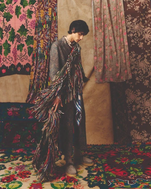 A young adult poses in a fashion editorial wearing a grey coat and pearl necklace, standing on a floral rug with patterned drapery behind.