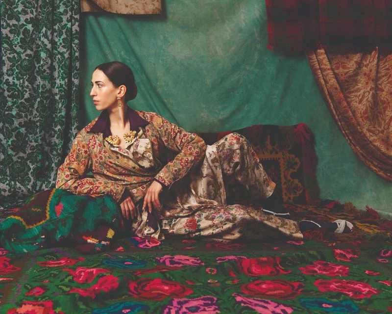 A woman with dark hair and a patterned jacket and pants lies on a floral carpet, with patterned drapes in the background, in an indoor setting.