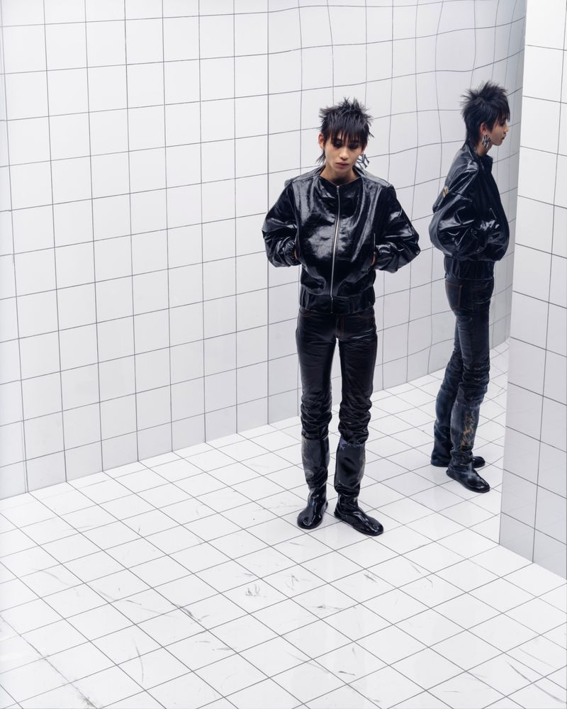 Fashion portrait of a young adult in shiny black jacket and pants standing in a corner with white square tiles and reflection.