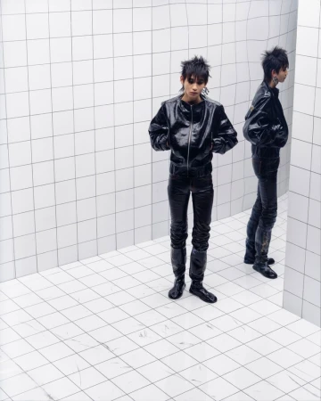Fashion portrait of a young adult in shiny black jacket and pants standing in a corner with white square tiles and reflection.
