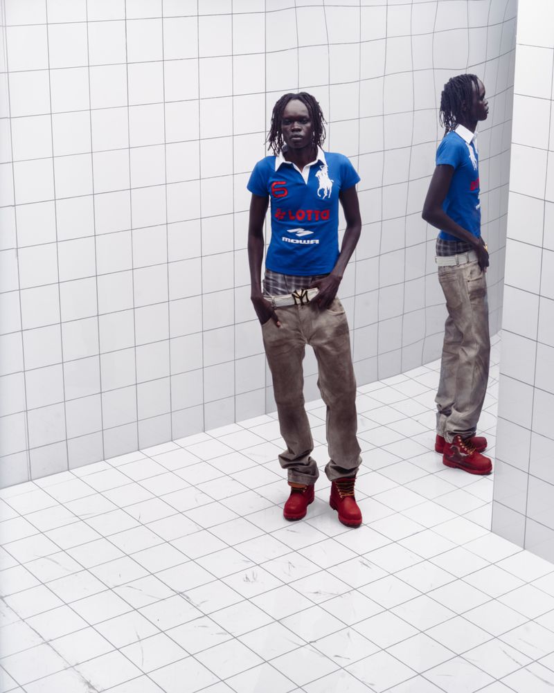 An African woman stands in a blue shirt, jeans, and red boots in a white-tiled room, her reflection visible in a mirror.