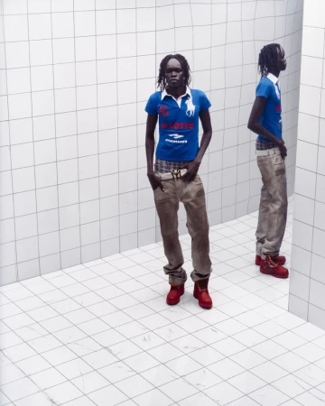An African woman stands in a blue shirt, jeans, and red boots in a white-tiled room, her reflection visible in a mirror.