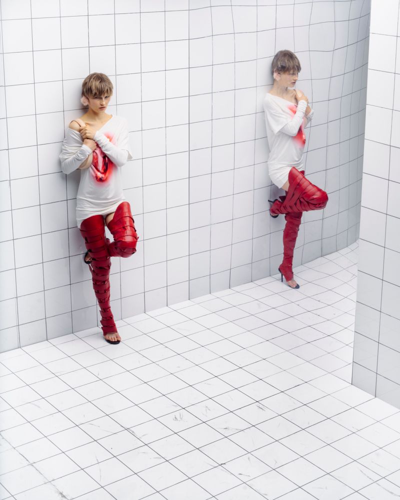 A young adult woman with short hair, wearing a white top and tall red leather boots, is posing in a white tiled room, fashion studio shot.