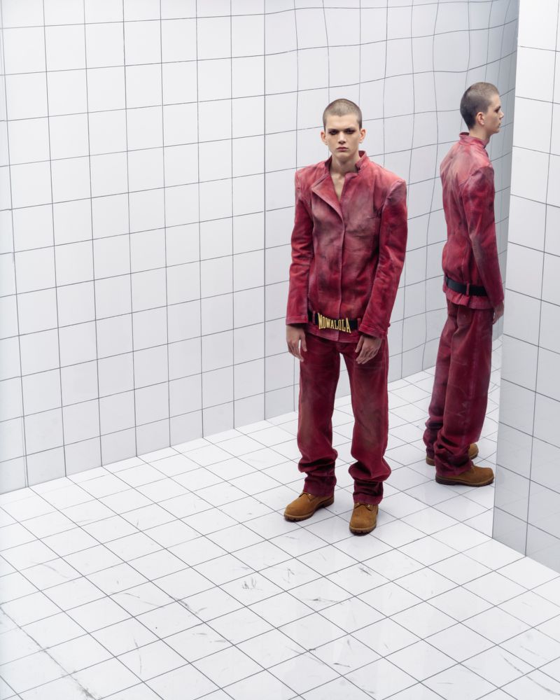 Young adult man in a matching red-burgundy jacket and pants, standing in a white tiled corner studio with a mirrored duplicate figure.