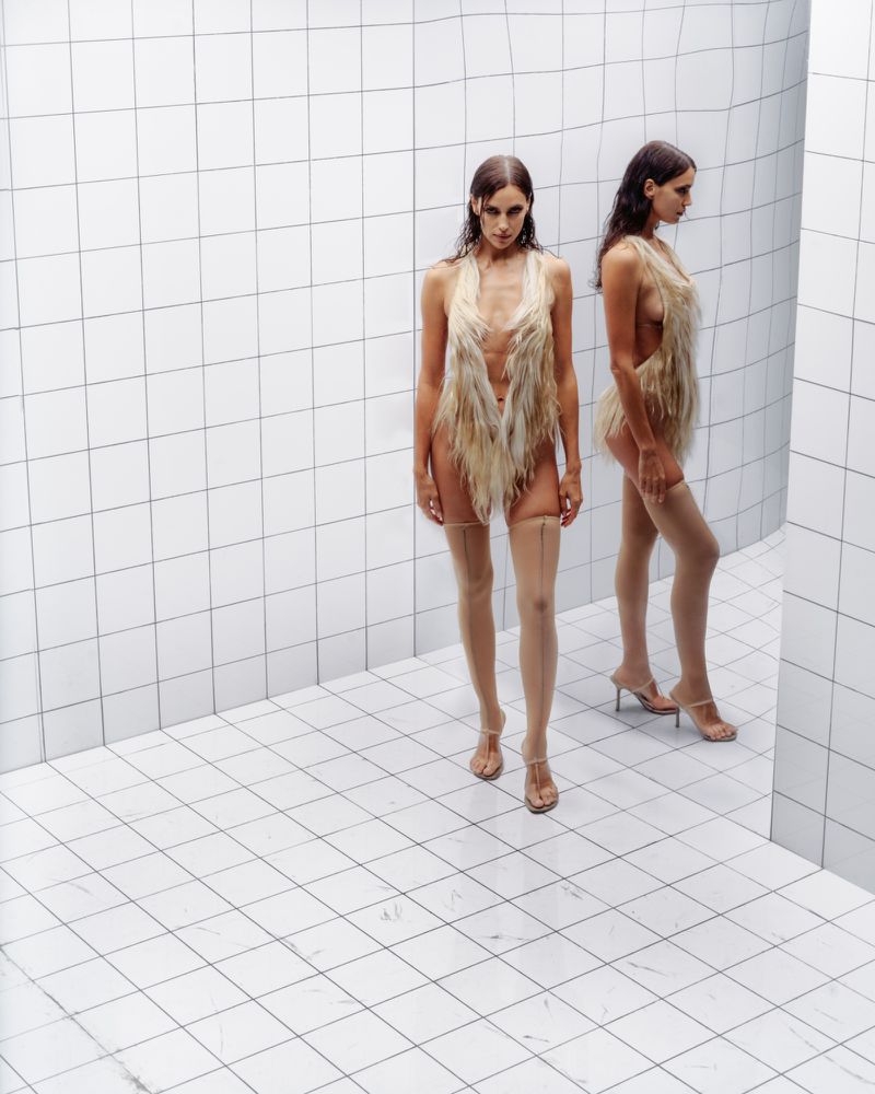 Two women in fur outfits and heels pose in a photo studio against a tiled wall and floor, creating a symmetrical, high-fashion image.