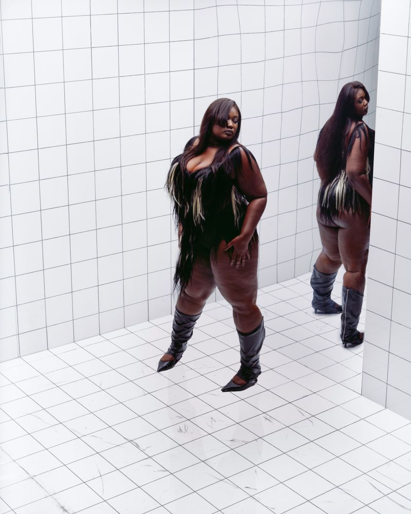 Full-length portrait of a Black woman wearing a dark fringed leotard and knee-high leather boots, posing in front of a white tiled background.