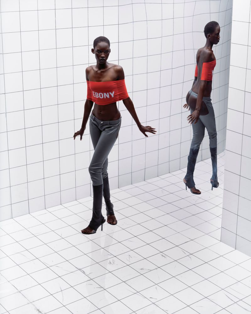 Two Black female fashion models pose in a photo studio wearing a red crop top, grey pants, and brown high-heeled boots against a tiled backdrop.