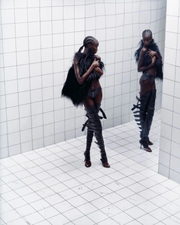 Two dark-skinned women model fashion in a white-tiled room. The women wear thigh-high strappy boots, underwear, and fur tops.