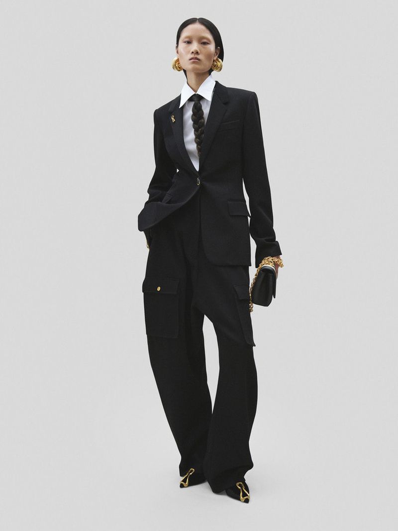 Asian woman in a black suit with a white shirt and tie poses in a studio with wide-leg pants, gold accessories, and black pointed heels.