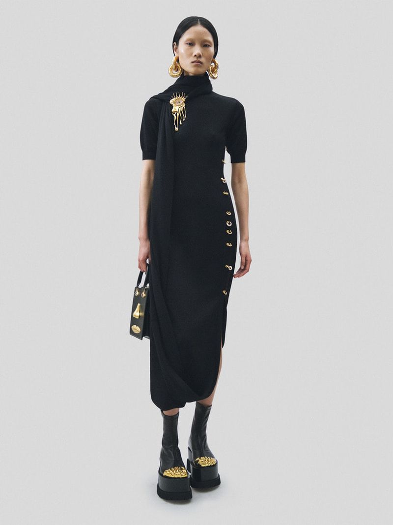 Adult woman in a black knit midi dress with gold button accents and platform shoes, accessorized with gold jewelry, stands in studio.