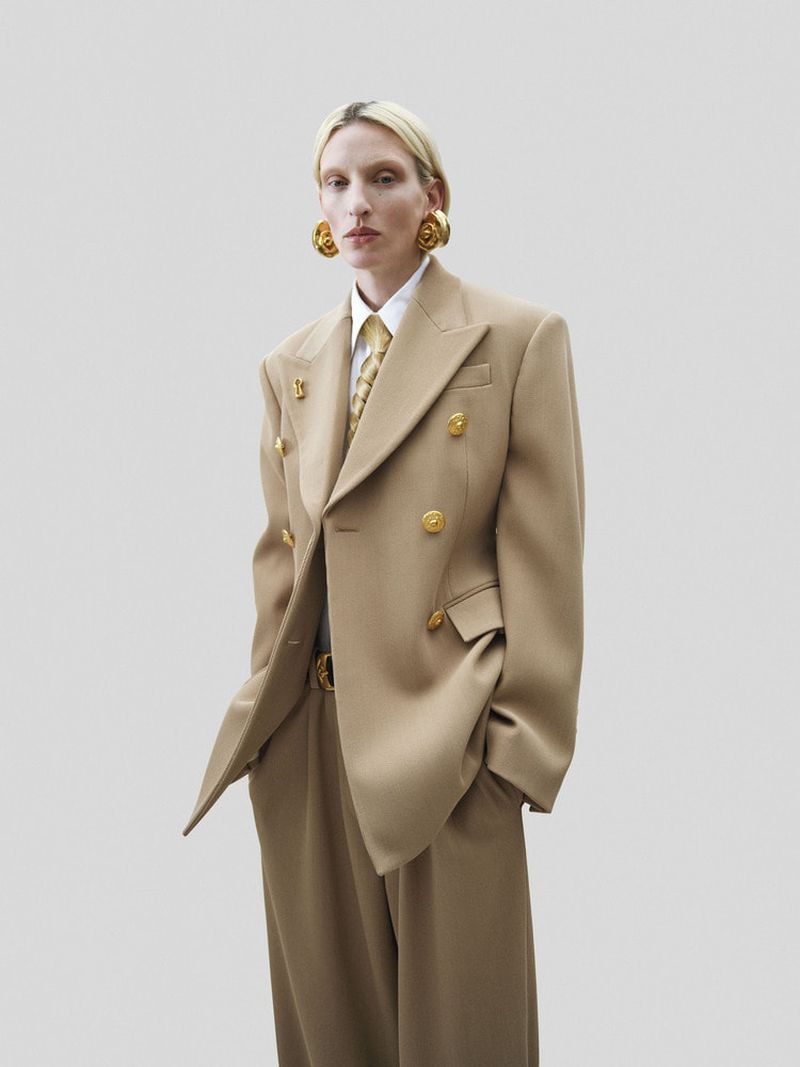 A fair-skinned woman with blonde hair, dressed in a tan double-breasted suit with gold buttons, a tie, and gold earrings, stands indoors.