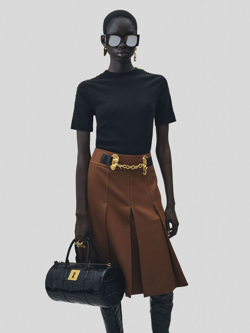 Model wearing black top, brown pleated skirt, and sunglasses, holding a black handbag against a grey background.
