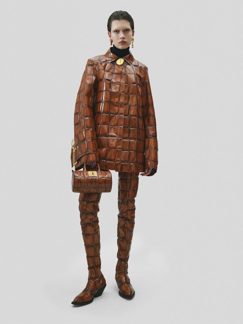 Model in glossy brown patterned leather coat and matching trousers holding a small handbag against a plain background