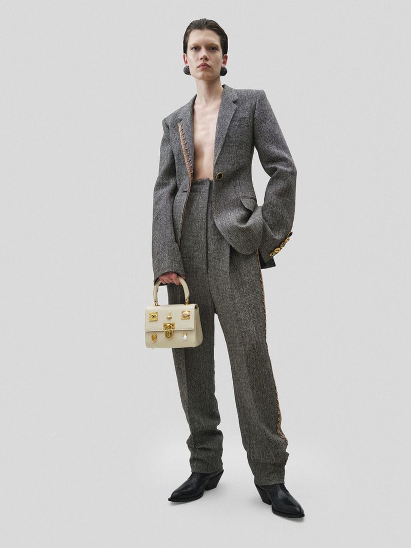 A model wears a grey tweed blazer and trousers with black heeled boots while holding a beige handbag in a studio shot.