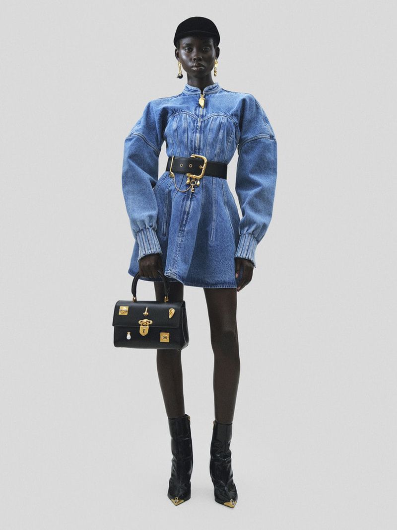 Fashionable Black woman poses in studio wearing denim mini dress with black belt, black handbag, and patent leather ankle boots.