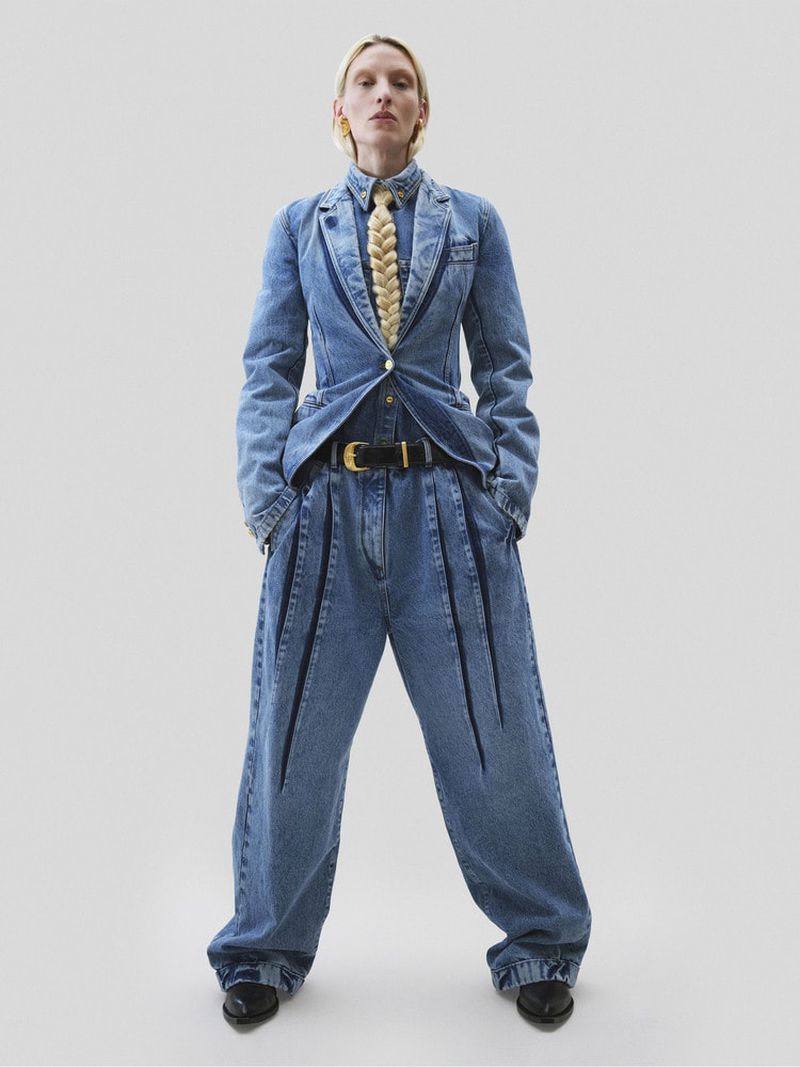A slender female fashion model poses in a light grey studio wearing a blue denim suit with tailored blazer and voluminous pleated jeans.