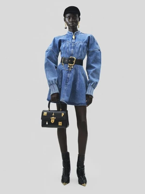 Model wearing a structured denim dress, black belt, and cap holding a black handbag against a neutral grey studio background.