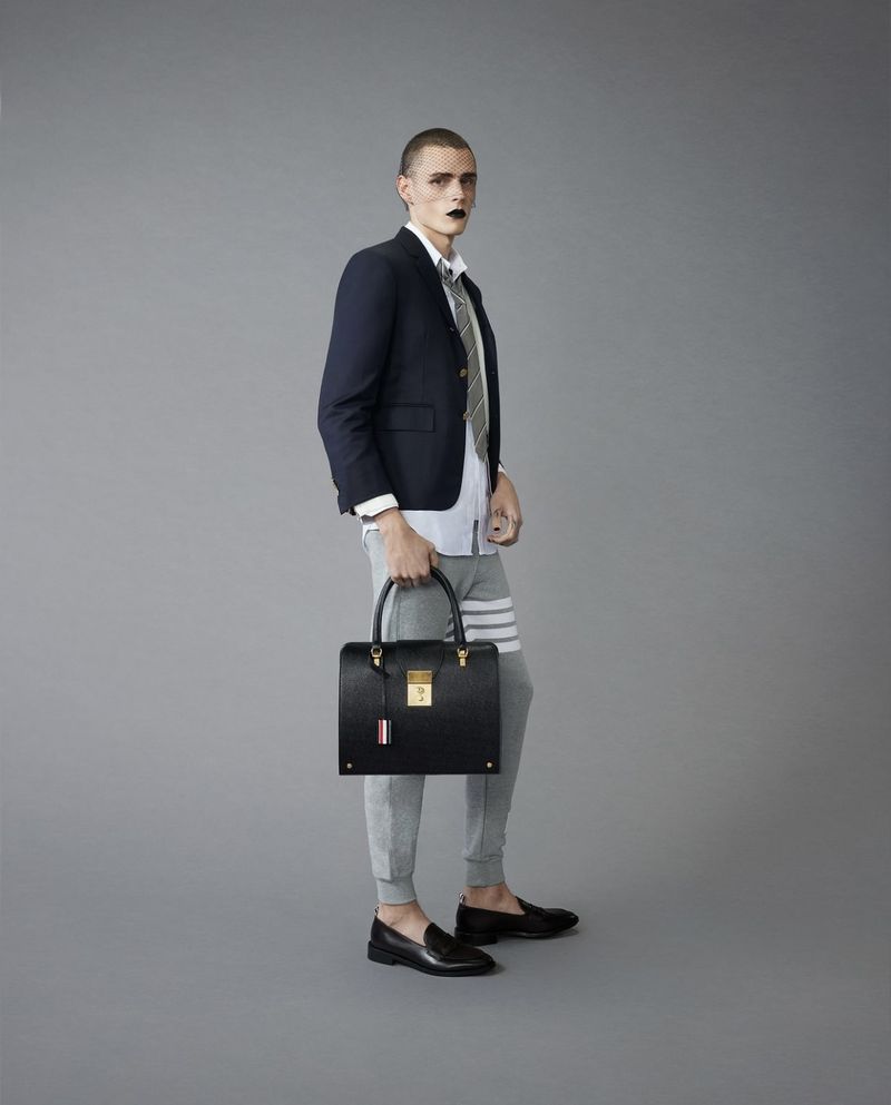 Male fashion model poses in grey sweatpants, loafers, navy blazer and tie while holding a black leather handbag in a studio shot.