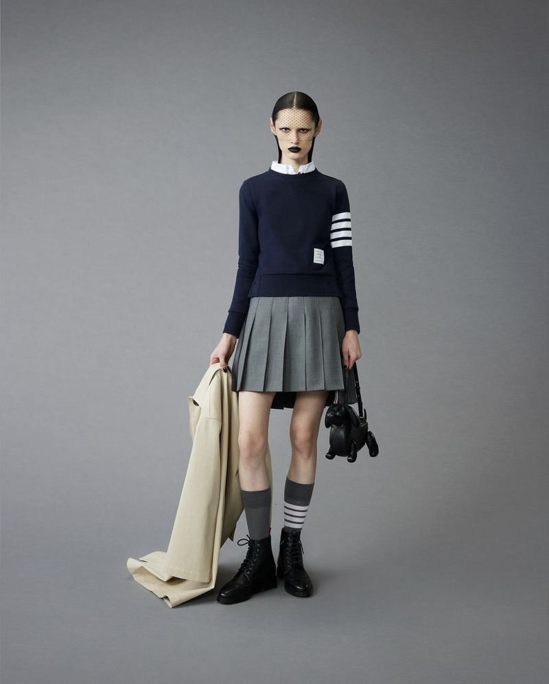 Fashion portrait of a woman in a navy sweater, pleated grey skirt, and black boots against a neutral grey background in studio lighting.