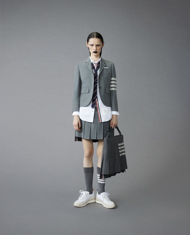 Fashion model in a Thom Browne school uniform with a grey blazer, pleated skirt, knee socks, and white sneakers, shot in a studio.