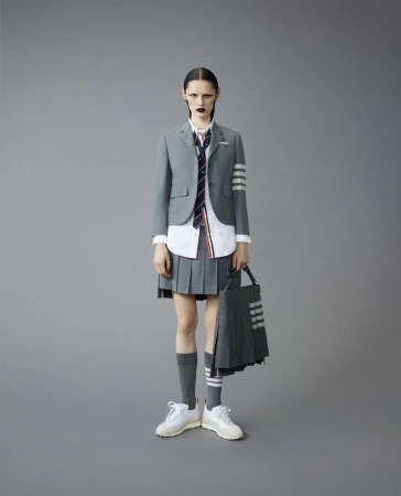 Fashion model in a Thom Browne school uniform with a grey blazer, pleated skirt, knee socks, and white sneakers, shot in a studio.