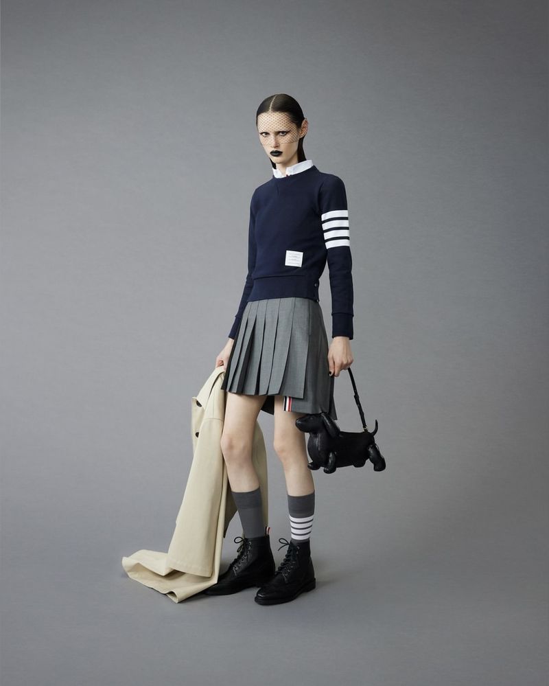 Fashion model poses in a navy sweater, grey pleated skirt, socks, and black boots, holding a beige coat and dog-shaped purse.