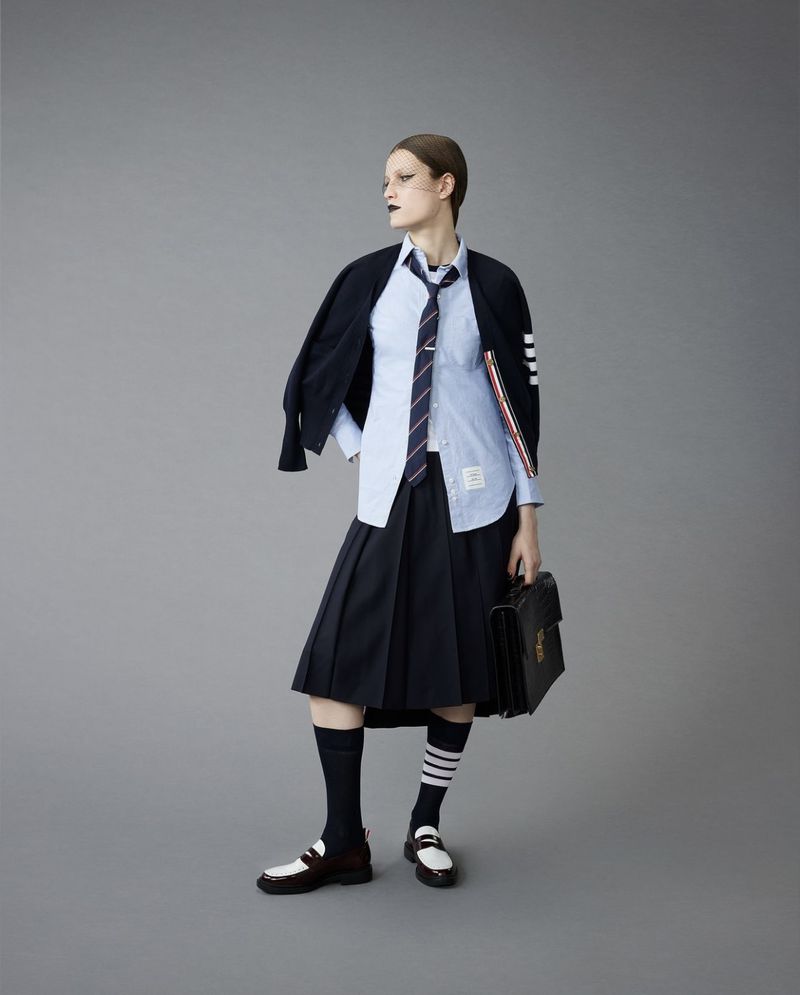 A woman poses in a preppy fashion look with a blue collared shirt, navy pleated skirt, loafers, knee socks, jacket, and briefcase.