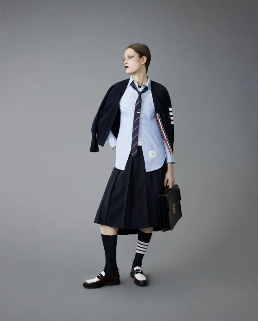 A woman poses in a preppy fashion look with a blue collared shirt, navy pleated skirt, loafers, knee socks, jacket, and briefcase.