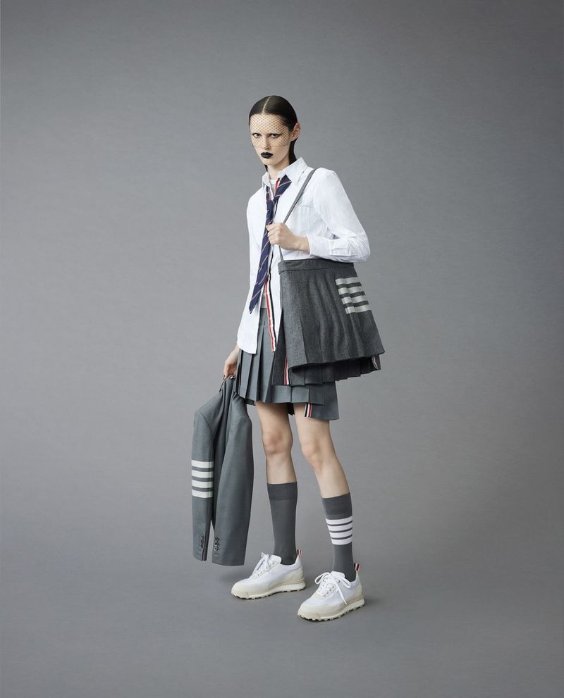 Fashion shot of a slender adult woman in a school uniform-inspired outfit, holding a blazer, set against a solid grey backdrop.
