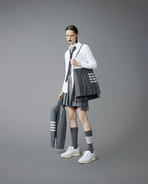 Fashion shot of a slender adult woman in a school uniform-inspired outfit, holding a blazer, set against a solid grey backdrop.