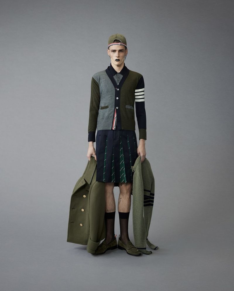 Male fashion model in modern menswear including cap, sweater, kilt, coat, and shoes, posing in a studio with a neutral grey background.