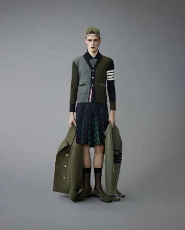 Male fashion model in modern menswear including cap, sweater, kilt, coat, and shoes, posing in a studio with a neutral grey background.
