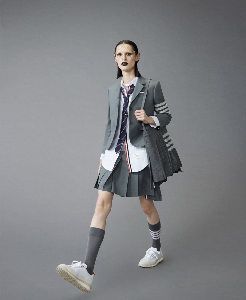 Fashion-forward adult woman strides confidently in grey blazer, pleated skirt, knee socks, and sneakers, set against a neutral grey background.