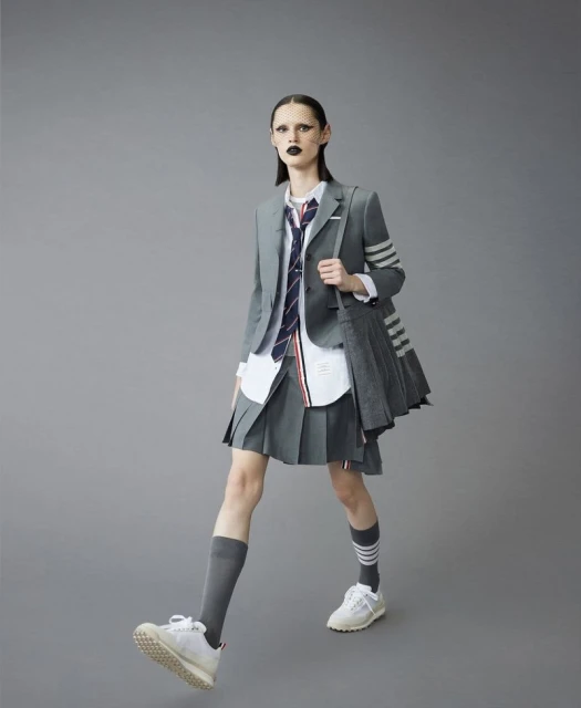 Fashion-forward adult woman strides confidently in grey blazer, pleated skirt, knee socks, and sneakers, set against a neutral grey background.