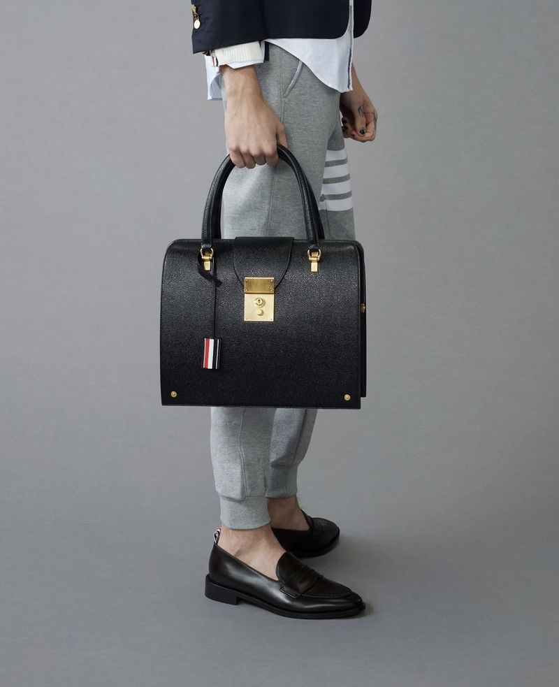 Person in a navy blazer, white shirt, grey sweatpants, and black loafers, holding a black leather handbag against a grey background.