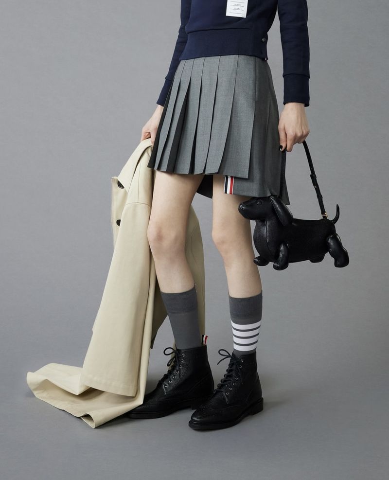 Fashionable young woman in pleated skirt, socks, boots, and dog-shaped purse. Light gray background. Studio lighting.